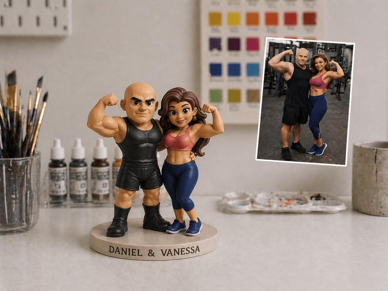 May include: A figurine of a muscular couple flexing their biceps. The male figure wears a black vest and shorts, while the female figure wears a pink sports bra and blue leggings. The base reads "DANIEL & VANESSA". A photo of the couple is in the background.