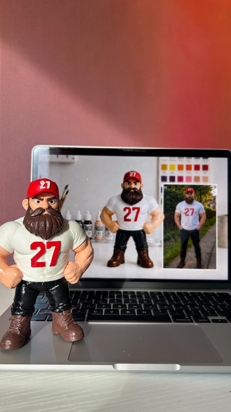May include: A figurine of a muscular man with a beard, wearing a white t-shirt with the number 27 in red, a red baseball cap, black trousers, and brown boots. The figurine is posed on a laptop, with images of the same figure on the screen.