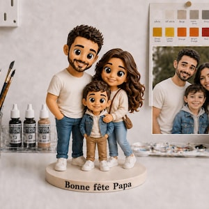 May include: A family figurine featuring a father, mother, and child in casual clothes, positioned on a round base. The base is inscribed with "Bonne f&ecirc;te Papa". A family photograph is in the background. Paintbrushes, paint bottles, and a colour palette are also visible.