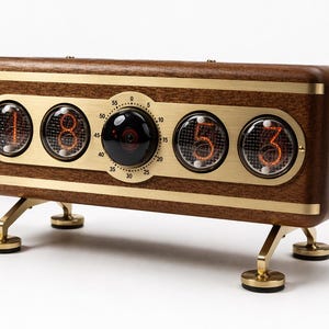 Puede incluir: Un reloj digital de estilo retro con marco de madera, detalles en latón y cuatro pantallas numéricas naranjas. El reloj tiene un dial central y está sostenido por patas de latón. La hora mostrada es 18:53.