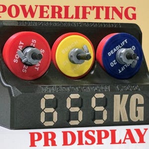 Peut inclure: Un présentoir de record personnel de powerlifting noir avec trois disques de poids rouges, jaunes et bleus. L'affichage indique "895 KG" en grands chiffres numériques, avec le mot "POWERLIFTING" au-dessus et "PR DISPLAY" en dessous.