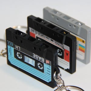 以下が含まれることがあります： 黒とグレーのカセットテープ型のキーホルダー3つ。一番手前のキーホルダーには「LH 60」と「BASE」の文字が入った青と黒のラベルが付いています。他の2つのキーホルダーには、赤、白、黄色のアクセントが付いたラベルが付いています。