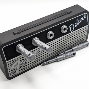 May include: A black and silver Fender Deluxe amplifier-shaped pen holder with two silver pens. The pen holder has a handle and the word "Deluxe" in white. The front has a silver grill design and the Fender logo.
