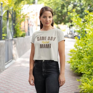 May include: A light beige crop top with the text "GAME DAY Football MAMI" in brown and gold lettering. The shirt is paired with black jeans. The image is taken outdoors with greenery in the background.