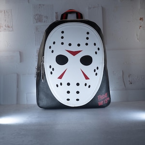 May include: A black backpack featuring a white hockey mask design on the front panel. The mask has black eye holes, red triangular markings, and black dots. The text FRIDAY THE 13TH is printed in red in the bottom right corner of the bag.