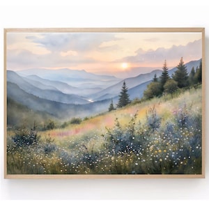 May include: Watercolor painting of a mountain landscape with a sunset. The artwork features layers of blue mountains, a grassy hillside with wildflowers, and evergreen trees. The painting is framed in a light-colored wooden frame.