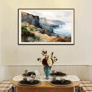 Puede incluir: Una pintura de acuarela enmarcada representa un paisaje costero con acantilados, oc&eacute;ano y un cielo brumoso. Debajo, una mesa de madera est&aacute; puesta con cubiertos, un jarr&oacute;n de flores y manteles individuales, creando una escena de comedor.
