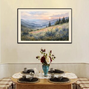 May include: A framed watercolor painting depicts a mountain landscape at sunset. The artwork features layers of blue mountains, a colorful meadow, and a vibrant sky. A dining table with place settings and a floral arrangement is in the foreground.