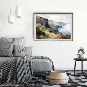 Puede incluir: Pintura de acuarela de un paisaje costero con acantilados, oc&eacute;ano y cielo. La obra de arte tiene un marco oscuro y se exhibe en una pared blanca sobre un sof&aacute; gris con almohadas y una manta a cuadros. Una cesta tejida est&aacute; en el suelo.
