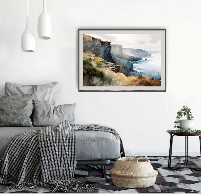 Puede incluir: Pintura de acuarela de un paisaje costero con acantilados, oc&eacute;ano y cielo. La obra de arte tiene un marco oscuro y se exhibe en una pared blanca sobre un sof&aacute; gris con almohadas y una manta a cuadros. Una cesta tejida est&aacute; en el suelo.