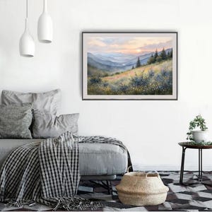 May include: A framed watercolor painting of a mountain landscape. The artwork depicts rolling hills, trees, and a sunset with shades of orange, yellow, and blue. The painting is displayed on a white wall above a gray sofa.
