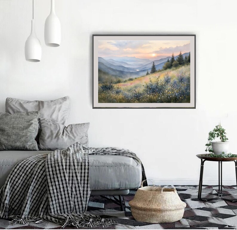 May include: A framed watercolor painting of a mountain landscape. The artwork depicts rolling hills, trees, and a sunset with shades of orange, yellow, and blue. The painting is displayed on a white wall above a gray sofa.