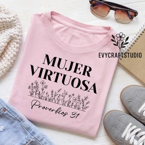 May include: A light pink t-shirt with the words "MUJER VIRTUOSA" and "Proverbios 31" in black script. A floral design is printed below the text. The shirt is displayed with sunglasses, a notebook, and gray sneakers.