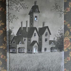 May include: A grayscale illustration of a spooky mansion with a ghost in a window. The book cover features the title "A Fraid of Ghosts" at the bottom, with a field of tall grass in the foreground and trees in the background.