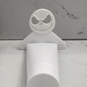 Puede incluir: Soporte de botella blanco con un diseño de cara de Jack Skellington. El soporte está diseñado para sujetar una botella cilíndrica. La parte superior del soporte tiene una cara con una sonrisa y ojos huecos.
