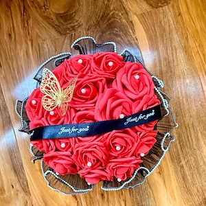 May include: A circular bouquet of red artificial roses, each featuring a small white pearl at its centre. The arrangement is accented with a gold-coloured filigree butterfly and wrapped in black mesh trim with white beads. A black ribbon reads Just for you.