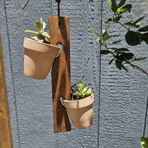 May include: A wooden hanging planter with two terracotta pots, each containing a small succulent plant. The dark brown wooden plank has heart-shaped cutouts to hold the pots. The background is a weathered, grey wooden fence.
