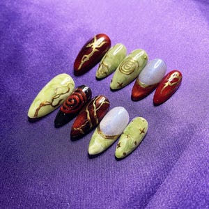 May include: A set of ten almond-shaped press-on nails featuring a variety of designs including deep red with gold filigree, marbled light green with gold swirls, iridescent white tips, and black with red spiral patterns and gold accents.