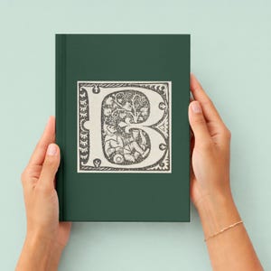 May include: A dark green journal with a decorative letter "B" on the cover. The letter is in a vintage style with an illustration of figures and foliage. The journal is held by two hands.