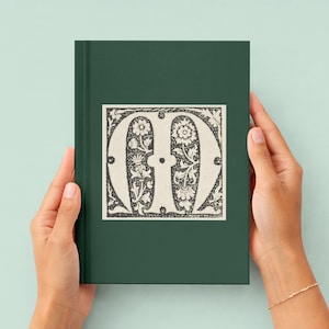May include: A dark green hardcover book featuring a central square illustration of a large, ornate, black and white letter M. The letter is decorated with intricate floral and botanical patterns, set against a solid dark green background.