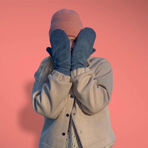 Puede incluir: Una persona con un gorro rosa claro, manoplas de pana azul y una chaqueta beige sobre un fondo coral. Las manoplas cubren la cara. La chaqueta tiene botones negros.