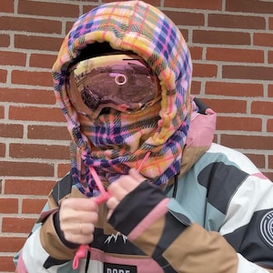 May include: A winter sports outfit featuring a plaid balaclava in pink, yellow, and blue, paired with mirrored goggles. The jacket has a color-block design with the word "DOPE" on the front.