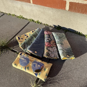 May include: A collection of colorful eyeglass cases and a pair of sunglasses. The cases feature various patterns, including a yellow geometric design, a blue quilted fabric, and floral prints. The sunglasses have silver frames and reflective lenses.