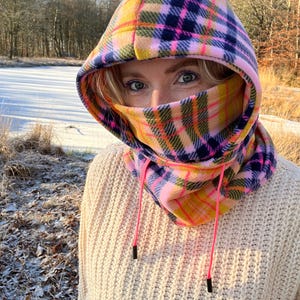 May include: A colourful plaid hooded scarf in pink, yellow, and navy blue. The scarf features a built-in face covering and adjustable pink drawstrings. The person is wearing a cream-coloured sweater.