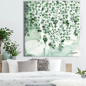 A bedroom with a white bed with a gray blanket and a wooden bench with a white faux fur throw. The bed is against a white wall with a large green and white abstract painting hanging above the bed. There are several potted plants in the room, including a large green plant on a wooden table next to the bed.