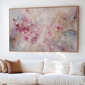 May include: Abstract floral painting in a gold frame. The artwork features a soft, blurred background with pink and white hues, and a cluster of bright pink, circular shapes. The painting hangs above a white sofa with brown cushions.