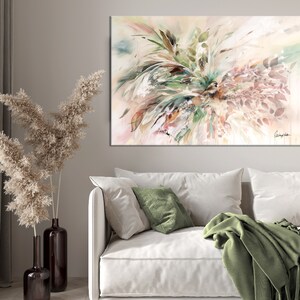 A large abstract painting with a white background and green, brown, and pink brushstrokes. The painting is hanging on a wall in a living room setting with a white couch, green throw blanket, and a vase with dried pampas grass.
