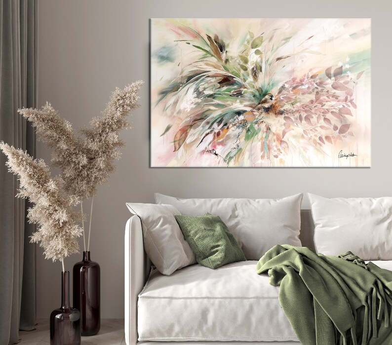 A large abstract painting with a white background and green, brown, and pink brushstrokes. The painting is hanging on a wall in a living room setting with a white couch, green throw blanket, and a vase with dried pampas grass.