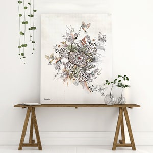 A black and white ink drawing of a floral design with birds and butterflies. The drawing is on a white canvas and is framed. The canvas is sitting on a wooden table with two small plants in glass vases.