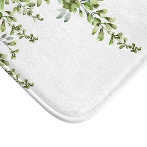 May include: White bath mat with a green watercolor floral design. The floral design is on the top edge of the mat and features leafy branches with small green flowers.