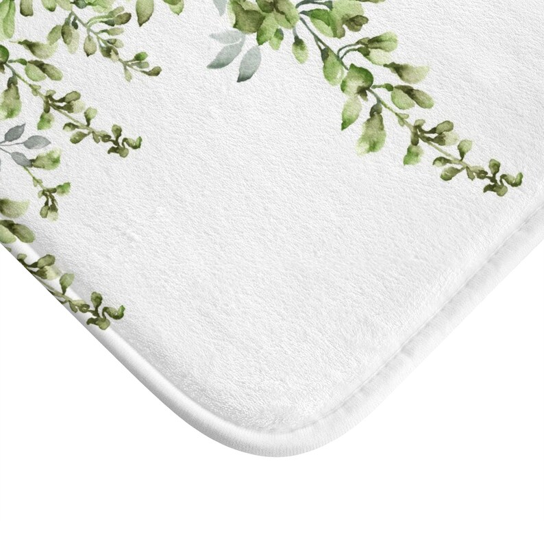 May include: White bath mat with a green watercolor floral design. The floral design is on the top edge of the mat and features leafy branches with small green flowers.