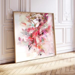 A framed floral art print featuring a vibrant display of pink, red, and white flowers. The artwork has a soft, watercolor-like effect, with a light background and a wooden frame. The print is leaning against a white wall.
