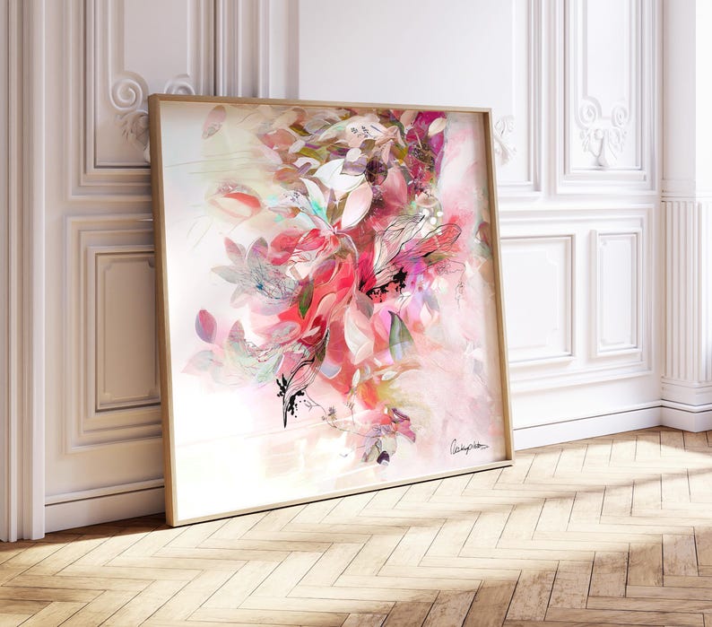 A framed floral art print featuring a vibrant display of pink, red, and white flowers. The artwork has a soft, watercolor-like effect, with a light background and a wooden frame. The print is leaning against a white wall.