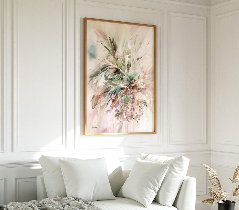 An abstract floral painting in a wooden frame. The artwork features a blend of soft pinks, greens, and beige hues, with brushstrokes creating a sense of movement and texture. The painting is displayed above a white sofa.