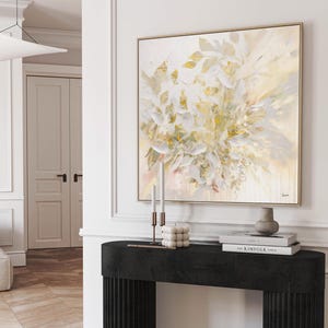 A square framed abstract painting with a floral design in shades of white, cream, and gold. The artwork hangs above a black console table with candles, books, and a vase. The room has white walls and a wooden floor.