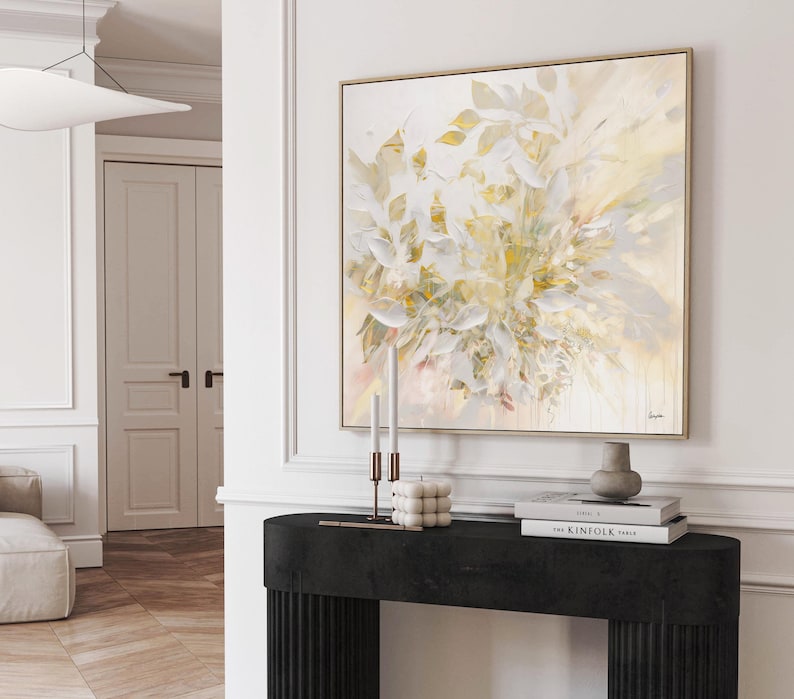 A square framed abstract painting with a floral design in shades of white, cream, and gold. The artwork hangs above a black console table with candles, books, and a vase. The room has white walls and a wooden floor.