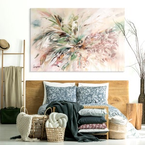 A bedroom interior with a wooden bed frame, a white wall, and a large abstract painting of pink, green, and white foliage. The bed is made with a blue and white patterned duvet cover, a dark green throw blanket, and a white knitted throw pillow. There are several woven baskets on the floor, and a wooden side table with a vase of dried branches.