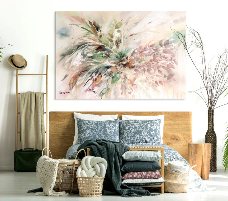 A bedroom interior with a wooden bed frame, a white wall, and a large abstract painting of pink, green, and white foliage. The bed is made with a blue and white patterned duvet cover, a dark green throw blanket, and a white knitted throw pillow. There are several woven baskets on the floor, and a wooden side table with a vase of dried branches.