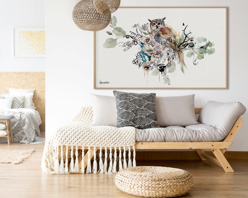 A watercolor painting of an owl perched on a branch with green leaves and flowers. The painting is framed and hanging on a white wall above a light gray couch with a white throw blanket.