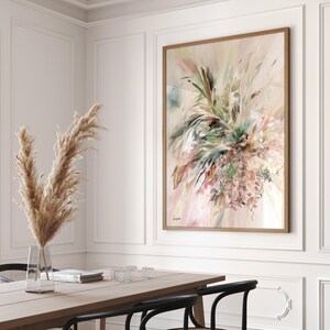A framed abstract watercolor painting with a wooden frame hangs on a white wall. The artwork features a blend of green, pink, and beige hues, depicting a floral or botanical theme. A vase of dried pampas grass sits on a wooden table.
