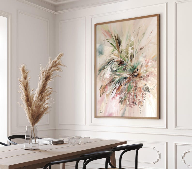 A framed abstract watercolor painting with a wooden frame hangs on a white wall. The artwork features a blend of green, pink, and beige hues, depicting a floral or botanical theme. A vase of dried pampas grass sits on a wooden table.