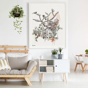 A watercolor-style illustration of a reindeer amidst flowers and birds, set against a beige background. The artwork is framed and displayed above a light beige futon with gold and beige pillows.  A white sideboard with books and plants sits below the artwork.  A hanging potted plant with trailing vines is visible in the upper left corner. The overall aesthetic is minimalist and nature-inspired.