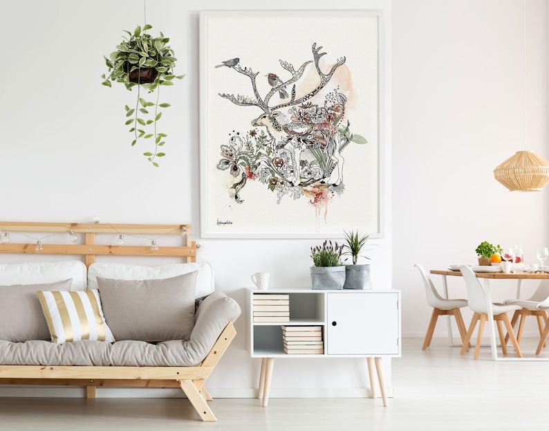 A watercolor-style illustration of a reindeer amidst flowers and birds, set against a beige background. The artwork is framed and displayed above a light beige futon with gold and beige pillows.  A white sideboard with books and plants sits below the artwork.  A hanging potted plant with trailing vines is visible in the upper left corner. The overall aesthetic is minimalist and nature-inspired.