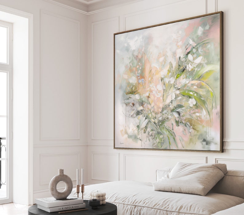 A large abstract painting with a light pink, green, and white color scheme. The painting depicts a cluster of flowers and leaves in a loose, impressionistic style. The painting is framed in a thin, gold-colored frame and is hanging on a white wall.