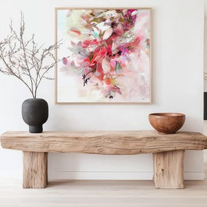 Abstract floral art print in a square wooden frame. The artwork features a vibrant composition of pink, red, and white brushstrokes, with hints of green and black accents. A dark gray vase with bare branches sits to the left, and a wooden bowl rests on a rustic wooden bench below the artwork.