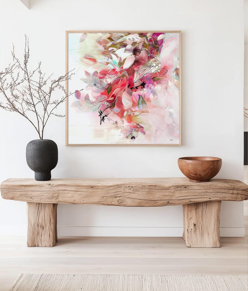 Abstract floral art print in a square wooden frame. The artwork features a vibrant composition of pink, red, and white brushstrokes, with hints of green and black accents. A dark gray vase with bare branches sits to the left, and a wooden bowl rests on a rustic wooden bench below the artwork.
